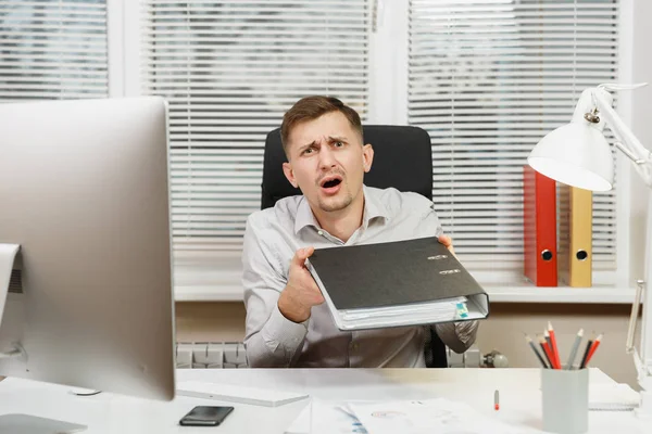 Шокированный и напряженный бизнесмен в рубашке сидит за столом, работает за компьютером с современным монитором, папками, лампой, документами в светлом кабинете на фоне окна. Менеджер или сотрудник . — стоковое фото