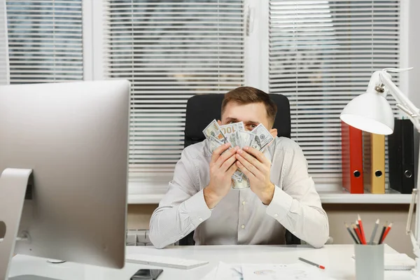 Όμορφος χαμογελαστός άνθρωπος των επιχειρήσεων στο πουκάμισο που κάθεται στο γραφείο του, κρύβεται πίσω από πολλά μετρητά χρήματα, εργάζονται σε υπολογιστή με σύγχρονη οθόνη, εγγράφων του office φως στο φόντο του παραθύρου. Διαχείριση ή εργαζόμενος. — Φωτογραφία Αρχείου