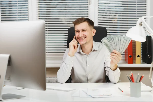Bilgisayarda çalışan modern monitör, lamba, ışık Office belgeleri ile yakışıklı gülümseyen iş tişörtlü adam çok nakit para, cep telefonuyla konuşurken masasında oturuyor. Yöneticisi veya alt.
