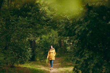 Güzel mutlu beyaz genç gülümseyen kahverengi saçlı kadın sarı ceket, kot, yeşil ormanda botlar. Ayakta ve erken sonbahar park açık havada yürüyüş sonbaharda altın yapraklarla kadın modeli moda.