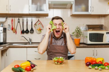 Eğlenceli deli beyaz genç adam önlük, kahverengi gömlek sebze salatası ile masada oturan yüz maskesi, ışık mutfakta gibi yeşil marul yaprağı koyun. Diyet kavramı. Evde yemek. Yemek hazırlamak.