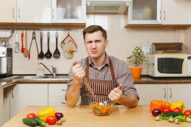 Çatal ve bıçak ışık mutfakta sebze salatası saldıran öfkeli deli aç genç adam masadan, önlük, kahverengi gömlekli. Diyet kavramı. Sağlıklı bir yaşam tarzı. Evde yemek. Yemek hazırlamak.
