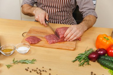 Domuz, sığır eti ya da kuzu eti hissesini ışık mutfak ahşap yüzeyli süslü mutfak eşyaları tam hazırlama evde yemek masada oturan sebze ile önlük beyaz genç adam kadar kapatın.
