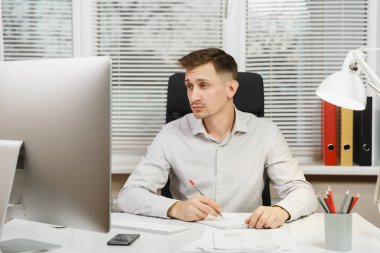 Pencere arka plan üzerinde ışık ofiste masa başında oturan gömlek ciddi ve dalmış iş adamı ile modern monitör, klasörler, kahve ya da çay, bilgisayarda çalışan makaleler. Yöneticisi veya alt.