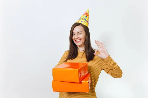 Bela diversão jovem feliz mulher em roupas amarelas, chapéu de festa de aniversário segurando caixas de presente laranja com presente, celebrando férias, mostrando gesto OK no fundo branco isolado para propaganda . — Fotografia de Stock