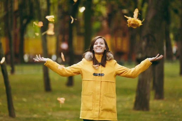 Piękny szczęśliwy kaukaski młody uśmiechający się brown-włos w żółtym fartuchu, dżinsy, buty w zielonym lesie. Modelka kobieta rzucając się spadek liści, stanie i chodzenie w początku jesieni na zewnątrz park. — Zdjęcie stockowe