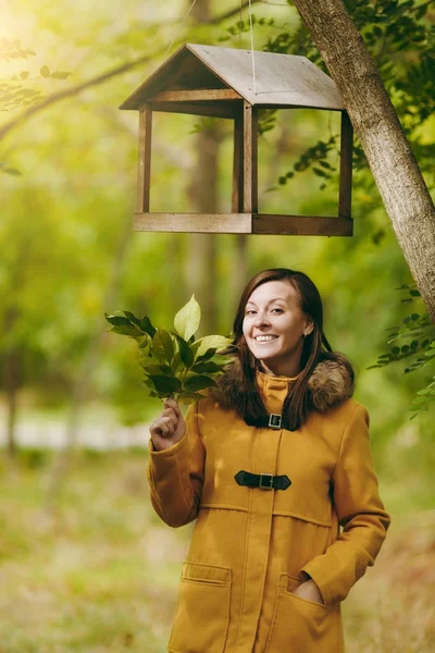 Piękny szczęśliwy kaukaski młody uśmiechający się brown-włos w żółtym fartuchu, dżinsy, buty w zielonym lesie. Modelka z jesieni pozostawia stojący pod karmniki w początku jesieni park na zewnątrz. — Zdjęcie stockowe