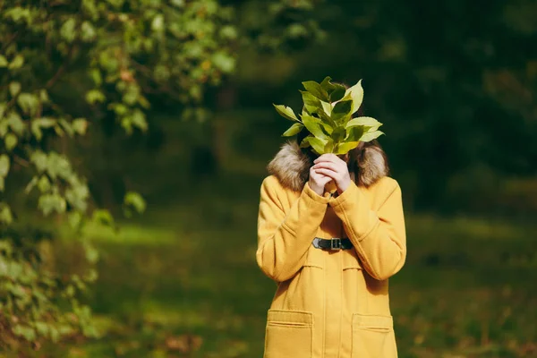 Hermosa mujer caucásica joven sonriente de pelo castaño en abrigo amarillo, jeans, botas en el bosque verde. Moda modelo femenino escondido detrás de las hojas de otoño de pie y caminando a principios de otoño parque al aire libre — Foto de Stock