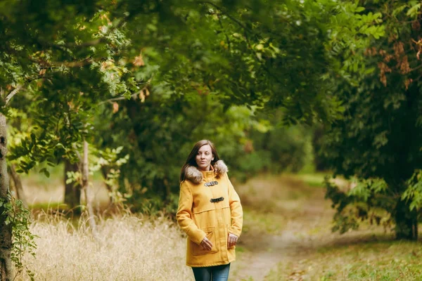 Piękny szczęśliwy kaukaski młody uśmiechający się brown-włos w żółtym fartuchu, dżinsy, buty w zielonym lesie. Mody modelki z liści jesienią złoty stanie i chodzenie w początku jesieni park na zewnątrz. — Zdjęcie stockowe