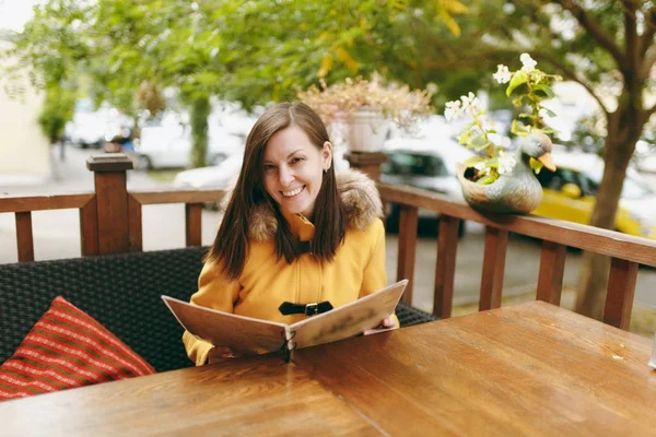 Piękny szczęśliwy kaukaski młody uśmiechający się brown-włos w żółtym fartuchu, czytanie, zamawianie z menu w restauracji lub kawiarni w pobliżu drogi i podejmowaniu decyzji, co jeść na kolacje w sezonie jesiennym. — Zdjęcie stockowe