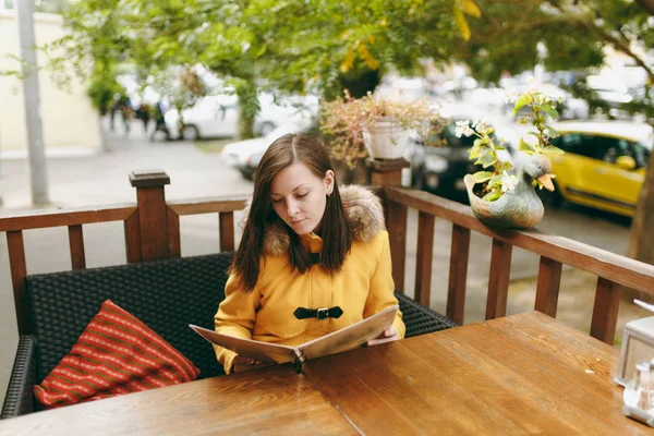 Schöne glückliche kaukasische junge lächelnde Frau mit braunem Haar in gelbem Mantel liest, bestellt von der Speisekarte im Außenrestaurant oder Café in der Nähe der Straße und entscheidet, was sie zum Abendessen im Herbst isst. — Stockfoto