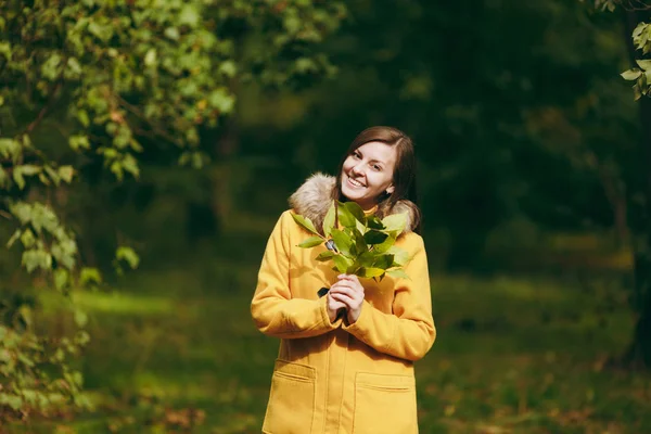 Beautiful happy caucasian young smiling brown-hair woman in yellow coat, jeans, boots in green forest. Fashion female model with fall golden leaves standing and walking in early autumn park outdoors. — Stock Photo, Image