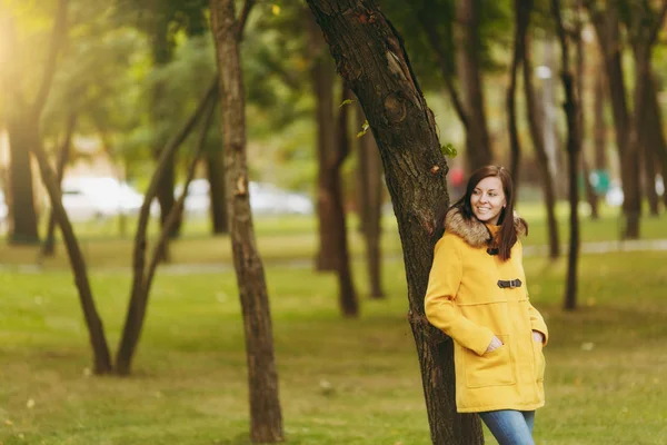 Piękny szczęśliwy kaukaski młody uśmiechający się brown-włos w żółtym fartuchu, dżinsy, buty w zielonym lesie. Mody modelki z liści jesienią złoty stanie i chodzenie w początku jesieni park na zewnątrz. — Zdjęcie stockowe