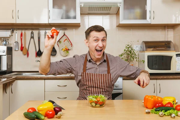 Fun jeune homme caucasien en tablier, chemise brune assise à table, choix entre oignon et tomate, coupe salade de légumes avec couteau dans la cuisine légère. Concept de régime. Cuisiner à la maison. Préparer les aliments . — Photo