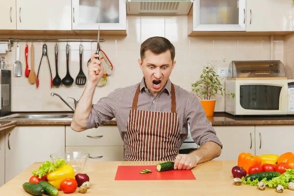 Zły szalony kaukaski młody człowiek w fartuch, brązowy shirt siedząc przy stole, atak kawałki warzyw na sałatkę z nożem w kuchni światło. Pojęcie diety. Zdrowy styl życia. Gotowanie w domu. Przygotowanie żywności. — Zdjęcie stockowe