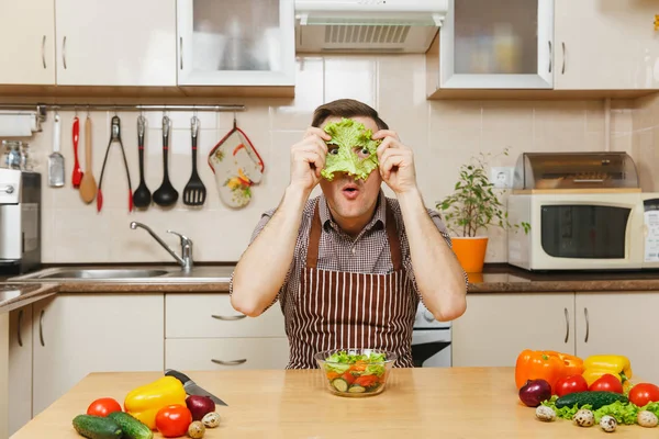 Веселый сумасшедший кавказский молодой человек в фартуке, коричневая рубашка сидит за столом с овощным салатом, кладет зеленый лист салата на лицо, как маска, на светлую кухню. Концепция диеты. Готовить дома. Готовьте еду. . — стоковое фото