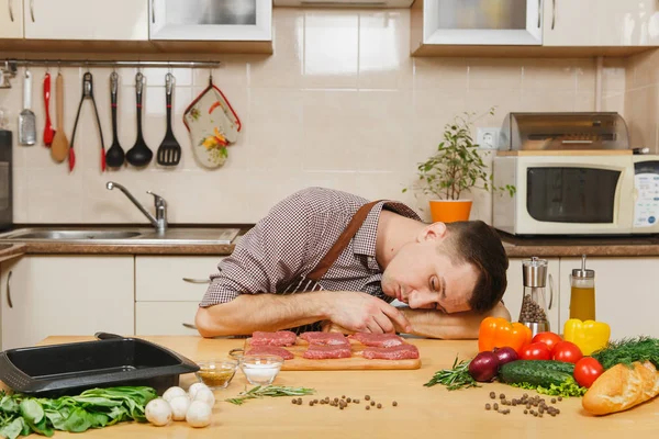 Caucasian young man in apron sleeping at table with vegetables, after cooking at home preparing meat stake from pork, beef or lamb, in light kitchen with wooden surface, full of fancy kitchenware.