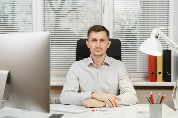 Pencere arka plan üzerinde ışık ofiste masa başında oturan gömlek ciddi ve dalmış iş adamı ile modern monitör, klasörler, kahve ya da çay, bilgisayarda çalışan makaleler. Yöneticisi veya alt.