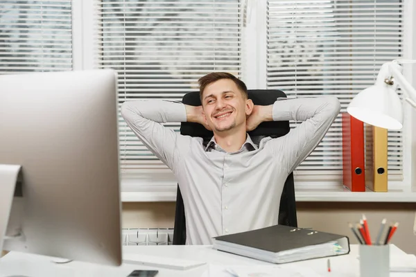 Schöner Geschäftsmann im Hemd sitzt am Schreibtisch, entspannt, hält die Hände hinter dem Kopf, arbeitet am Computer mit modernem Monitor, Dokumente im hellen Büro auf Fensterhintergrund. Manager oder Arbeiter. — Stockfoto