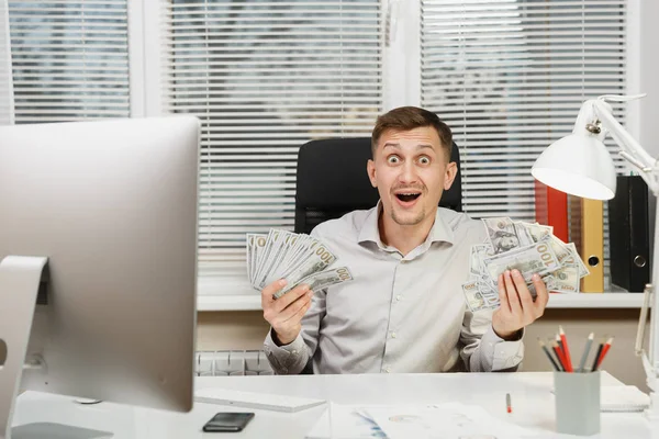 Ευτυχισμένος χαμογελαστός άνθρωπος των επιχειρήσεων στο πουκάμισο που κάθεται στο γραφείο του με πολλά μετρητά χρήματα, εργάζονται σε υπολογιστή με σύγχρονη οθόνη, εγγράφων του office φως στο φόντο του παραθύρου. Διαχείριση ή εργαζόμενος νικητής. — Φωτογραφία Αρχείου