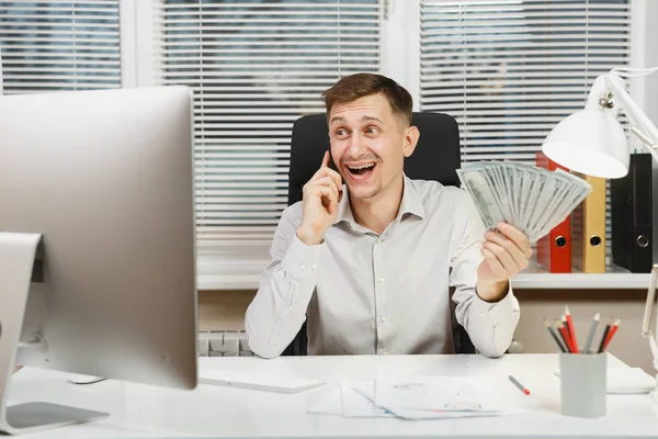 Beau homme d'affaires souriant en chemise assis au bureau parlant sur un téléphone mobile avec beaucoup d'argent comptant, travaillant à l'ordinateur avec moniteur moderne, lampe, documents dans le bureau léger. Directeur ou travailleur . — Photo