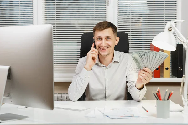 Όμορφος χαμογελαστός άνθρωπος των επιχειρήσεων στο πουκάμισο που κάθεται στο γραφείο του, μιλάμε για κινητό τηλέφωνο με πολλά μετρητά χρήματα, εργάζονται σε υπολογιστή με σύγχρονο monitor, λάμπα, εγγράφων του office φως. Διαχείριση ή εργαζόμενος. — Φωτογραφία Αρχείου