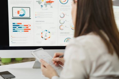 Masa başında oturan takım elbiseli Brown-saç iş kadını kalem bilgi ile defter için bilgisayarda çerçevede modern monitör, geri ışık Office belgeleri ile çalışan görünüm yazma