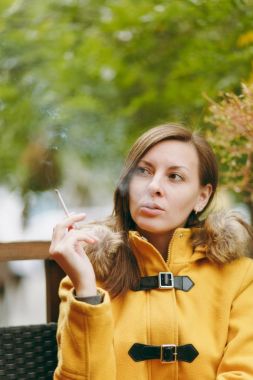 Güzel mutlu beyaz genç gülümseyen kahverengi saçlı kadın sarı ceket duman sigara açık restoranda veya kafe Sonbahar sezonu. Dinlenme, duman, sakin ol.