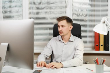 Bilgisayar başında modern monitör ile çalışan yakışıklı gülümseyen iş tişörtlü adam masada oturan eller klavye, klasörler, belge penceresi arka plan üzerinde ışık ofiste. Yöneticisi veya alt.