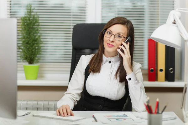 Beautiful smiling business woman in suit and glasses sitting at the desk, working at contemporary computer with documents in light office, talking on mobile phone, conducting pleasant conversation — Stock Photo, Image