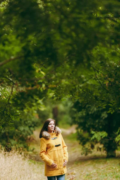 Bonito feliz caucasiano jovem sorrindo mulher de cabelo castanho em casaco amarelo, jeans, botas na floresta verde. Moda modelo feminino com outono folhas douradas de pé e andando no início do outono parque ao ar livre . — Fotografia de Stock