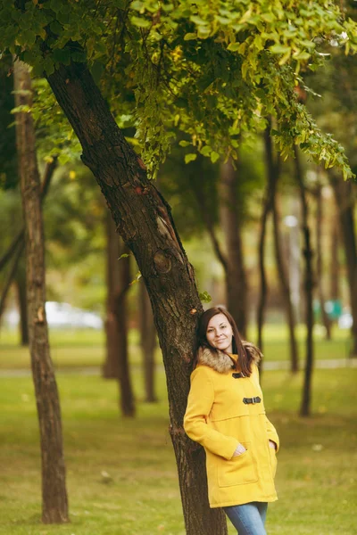 Vackra glada kaukasiska unga leende brown-hår kvinna i gul jacka, jeans, boots i gröna skogen. Mode kvinnlig modell med fall gyllene blad stående och gående i tidig höst park utomhus. — Stockfoto