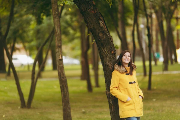 Красивая счастливая кавказская молодая улыбающаяся каштановая женщина в желтом пальто, джинсах, сапогах в зеленом лесу. Мода женская модель с осенью золотые листья стоят и ходить в ранней осени парк на открытом воздухе . — стоковое фото