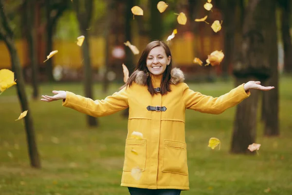 Beautiful happy caucasian young smiling brown-hair woman in yellow coat, jeans, boots in green forest. Fashion female model throwing up fall leaves standing and walking in early autumn park outdoors.