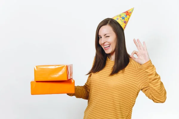 Bela diversão jovem feliz mulher em roupas amarelas, chapéu de festa de aniversário segurando caixas de presente laranja com presente, celebrando férias, mostrando gesto OK no fundo branco isolado para propaganda . — Fotografia de Stock
