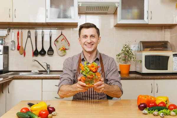 Красивый веселый кавказский молодой человек в фартуке, коричневая рубашка сидит за столом, блюет овощной салат в миске на светлой современной кухне. Концепция диеты. Здоровый образ жизни. Готовить дома. Готовьте еду. . — стоковое фото
