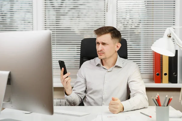 Serious business man in shirt sitting at the desk, talking on mobile phone, resolving issues, working at computer with modern monitor, documents in light office on window background. Manager or worker — Stock Photo, Image