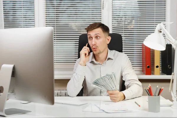 Όμορφος χαμογελαστός άνθρωπος των επιχειρήσεων στο πουκάμισο που κάθεται στο γραφείο του, μιλάμε για κινητό τηλέφωνο με πολλά μετρητά χρήματα, εργάζονται σε υπολογιστή με σύγχρονο monitor, λάμπα, εγγράφων του office φως. Διαχείριση ή εργαζόμενος. — Φωτογραφία Αρχείου