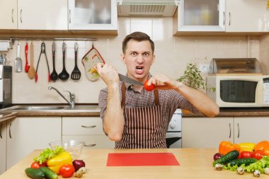 Öfkeli deli beyaz genç adam masada oturan önlük, kahverengi gömlekli saldırı sebze salatası için ışık mutfakta bıçakla keser. Diyet kavramı. Sağlıklı bir yaşam tarzı. Evde yemek. Yemek hazırlamak.