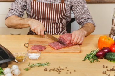 Domuz, sığır eti ya da kuzu eti hissesini ışık mutfak ahşap yüzeyli süslü mutfak eşyaları tam hazırlama evde yemek masada oturan sebze ile önlük beyaz genç adam kadar kapatın.