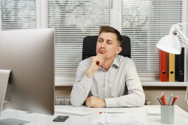 Pencere arka plan üzerinde ışık ofiste masa başında oturan gömlek ciddi ve dalmış iş adamı ile modern monitör, klasörler, kahve ya da çay, bilgisayarda çalışan makaleler. Yöneticisi veya alt.