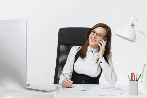 Pretty smiling business woman in suit sitting at the desk, working at contemporary computer with document in light office, talking on mobile phone, conducting pleasant conversation on white background — Stock Photo, Image