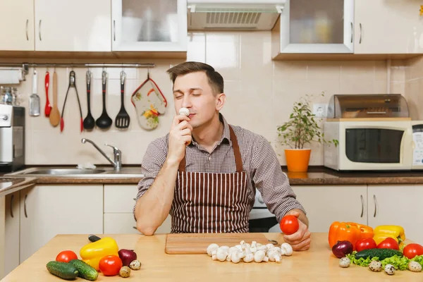 Красивый кавказский молодой человек в фартуке, коричневая рубашка сидит за столом с овощами, режет шампиньоны на светлой кухне. Концепция диеты. Здоровый образ жизни. Готовить дома. Готовьте еду. . — стоковое фото