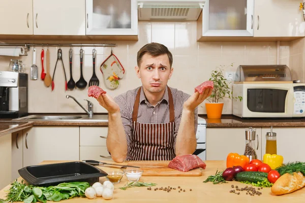 Trauriger aufgebrachter kaukasischer junger Mann in Schürze, der am Tisch mit Gemüse sitzt und zu Hause Fleisch vom Schwein, Rind oder Lamm zubereitet, in heller Küche mit Holzoberfläche, voller schickem Geschirr. — Stockfoto