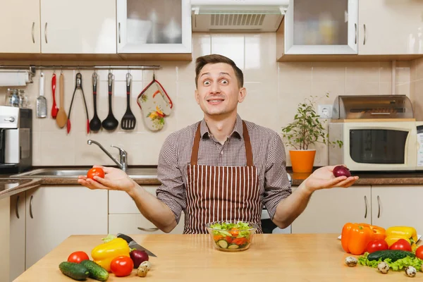 Ein lustiger kaukasischer junger Mann in Schürze, braunem Hemd sitzt am Tisch, wählt zwischen Zwiebeln und Tomaten, schneidet Gemüsesalat mit Messer in der hellen Küche. Diätkonzept. Kochen zu Hause. Essen zubereiten. — Stockfoto