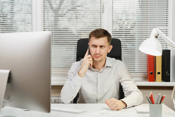 Hombre de negocios serio con camisa sentado en el escritorio, hablando por teléfono móvil, resolviendo problemas, trabajando en la computadora con monitor moderno, documentos en la oficina de luz en el fondo de la ventana. Gerente o trabajador — Foto de Stock