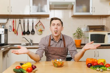 Kase ve Bıldırcın yumurta içinde ağız içinde sebze salatası ile masada oturmuş, hafif mutfak elinde yayılan önlük, kahverengi gömlekli eğlenceli beyaz genç adam. Diyet kavramı. Evde yemek. Yemek hazırlamak.