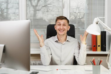 Eller, bilgisayarda çalışan modern monitör, lamba, klasörler, belge penceresi arka plan üzerinde ışık Office ile yayılan yakışıklı gülümseyen iş tişörtlü adam masada oturuyor. Yöneticisi veya alt.