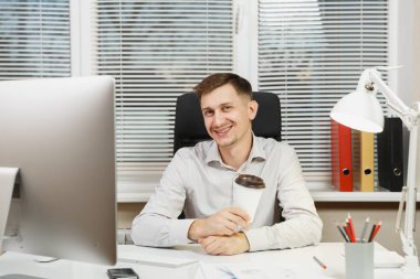 Modern İzleyicisi ile bilgisayar, belge penceresi arka plan üzerinde ışık ofiste çalışan yakışıklı gülümseyen iş tişörtlü adam fincan kahve ya da çay, ile masaya oturuyor. Yöneticisi veya alt.