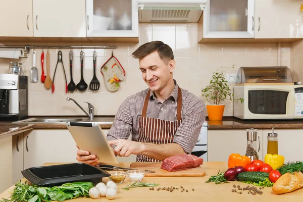 Junger Mann in Schürze sitzt am Tisch mit Gemüse, sucht Rezept in Tablette, kocht zu Hause Fleischpfahl aus Rindfleisch, in heller Küche mit Holzoberfläche, voller schicker Küchenutensilien. — Stockfoto